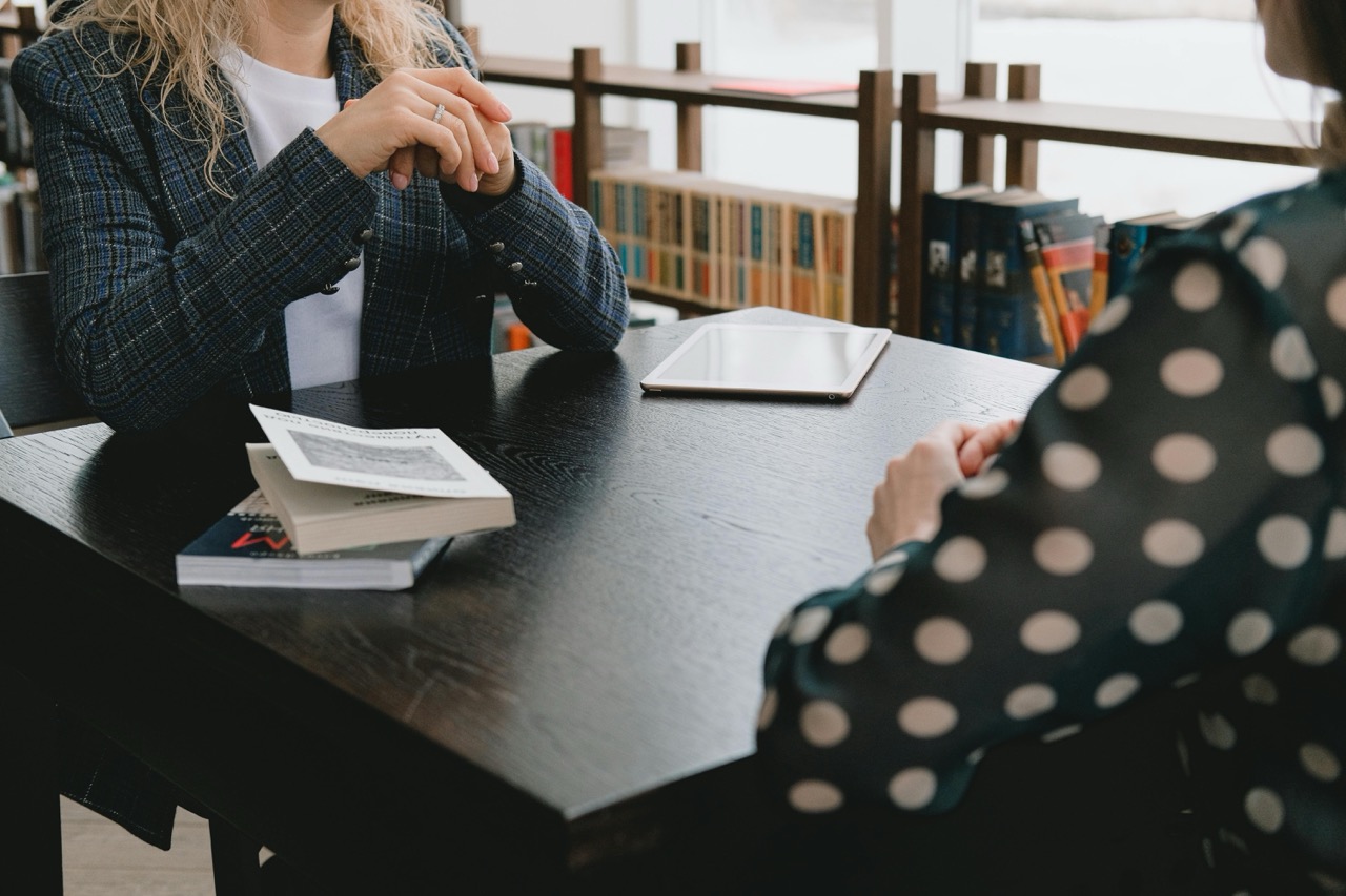 Dwie osoby siedzą przy stole i rozmawiają. Na stole leżą książki oraz tablet. W tle widoczna jest półka z książkami. Kadr pokazuje dłonie i fragmenty sylwetek rozmówców.
