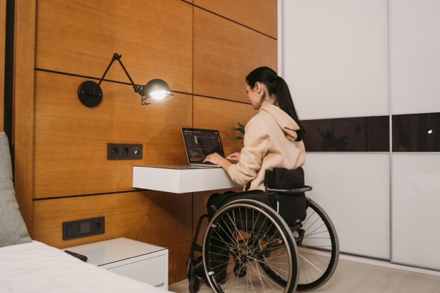 Kobieta na wózku inwalidzkim pracuje przy laptopie na biurku przytwierdzonym do ściany w nowocześnie urządzonym pokoju. Ma na sobie beżową bluzę z kapturem i siedzi przodem do biurka. Na ścianie obok znajduje się nowoczesna lampa oraz gniazdka elektryczne.