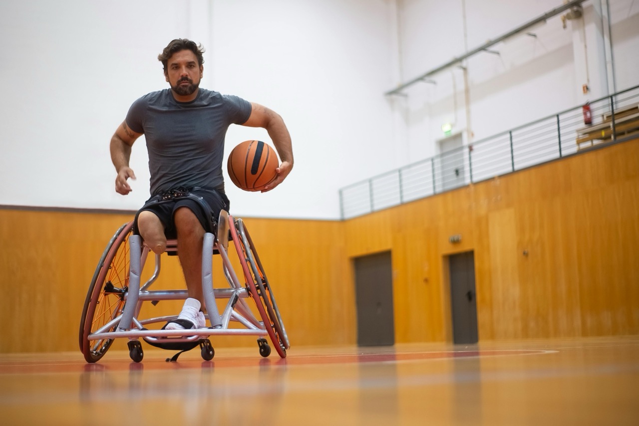 Wysportowany mężczyzna z jedną kończyną dolną, na wózku, gra w koszykówkę na hali sportowej – dynamiczne ujęcie pokazujące siłę, determinację i aktywność.