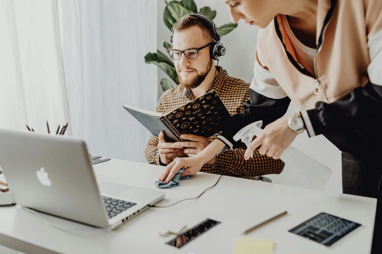 Biuro w jasnym świetle dziennym: mężczyzna w okularach i słuchawkach siedzi przy biurku, trzyma otwarty notes i spogląda na ekran laptopa, podczas gdy obok druga osoba spryskuje i wyciera blat miękką ściereczką; na stole leżą telefon, klawiatura i długopis, w tle roślina doniczkowa - scena łącząca pracę z dbałością o czystość stanowiska.