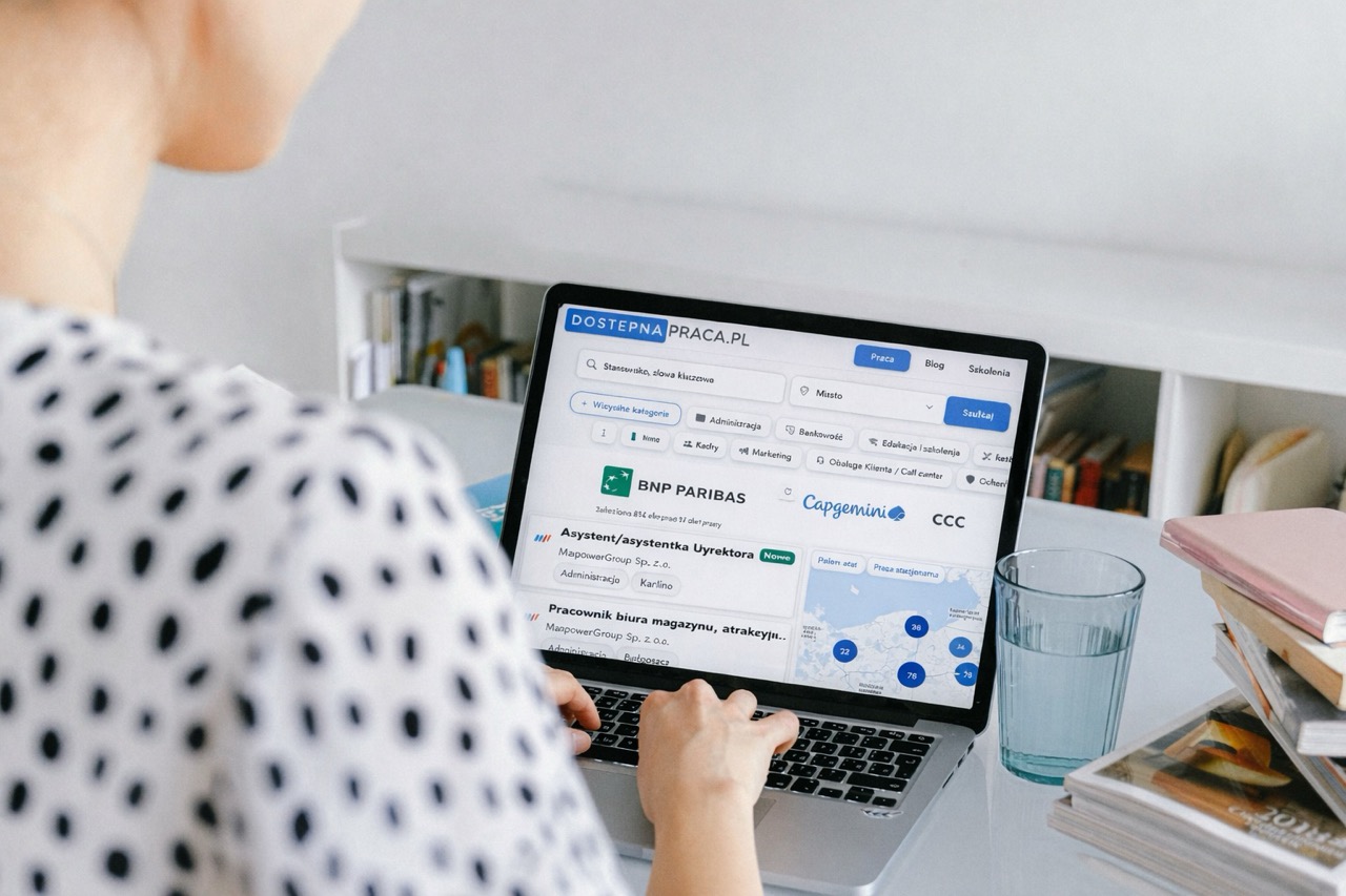 Osoba siedzi przy biurku i korzysta z laptopa. Na ekranie widoczna jest strona internetowa z ofertami pracy. Obok laptopa stoi szklanka wody i leżą książki.